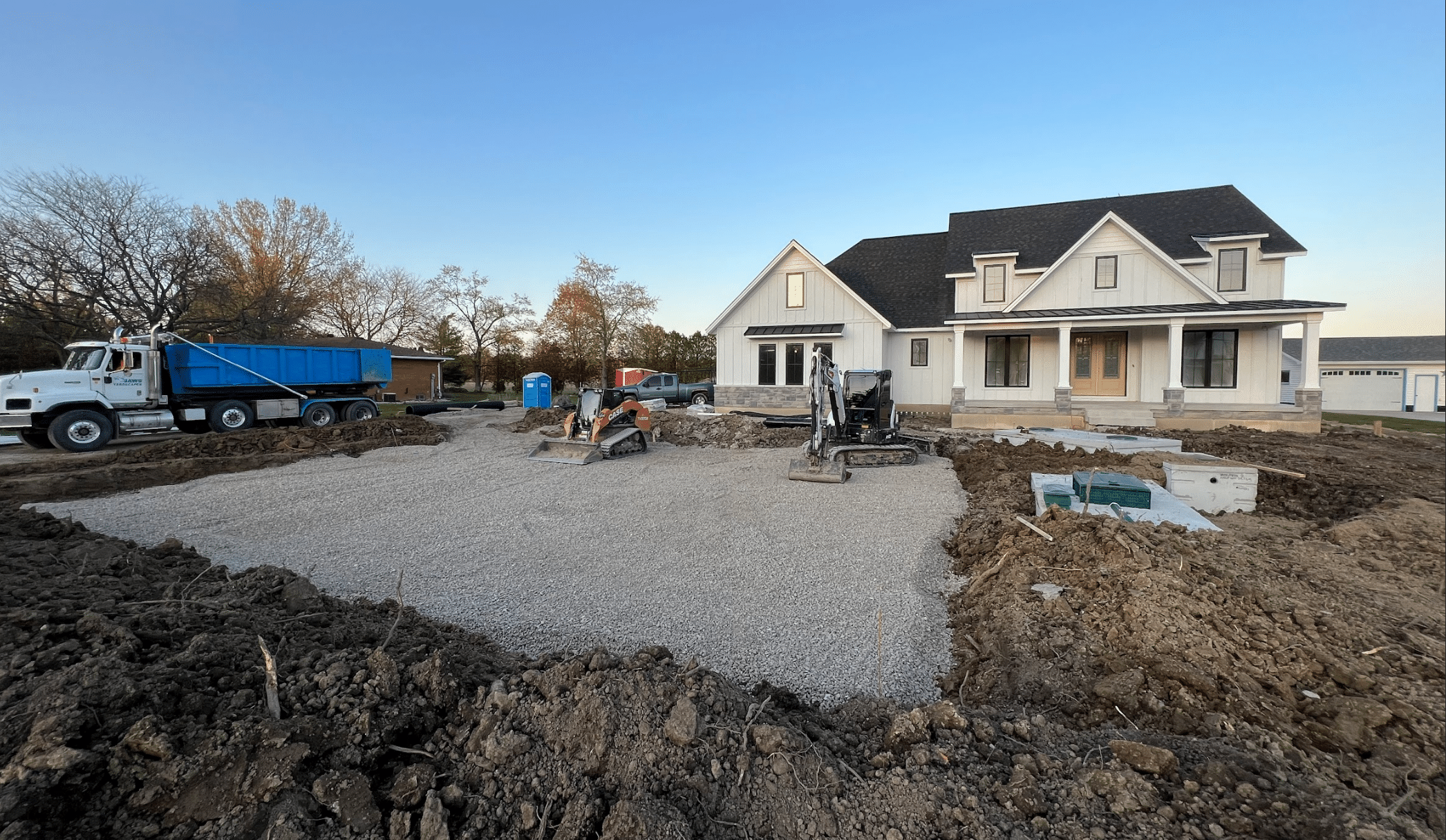Construction equipment and a truck are in front of a house with a gravel-covered yard, indicating ongoing landscaping or driveway installation work.