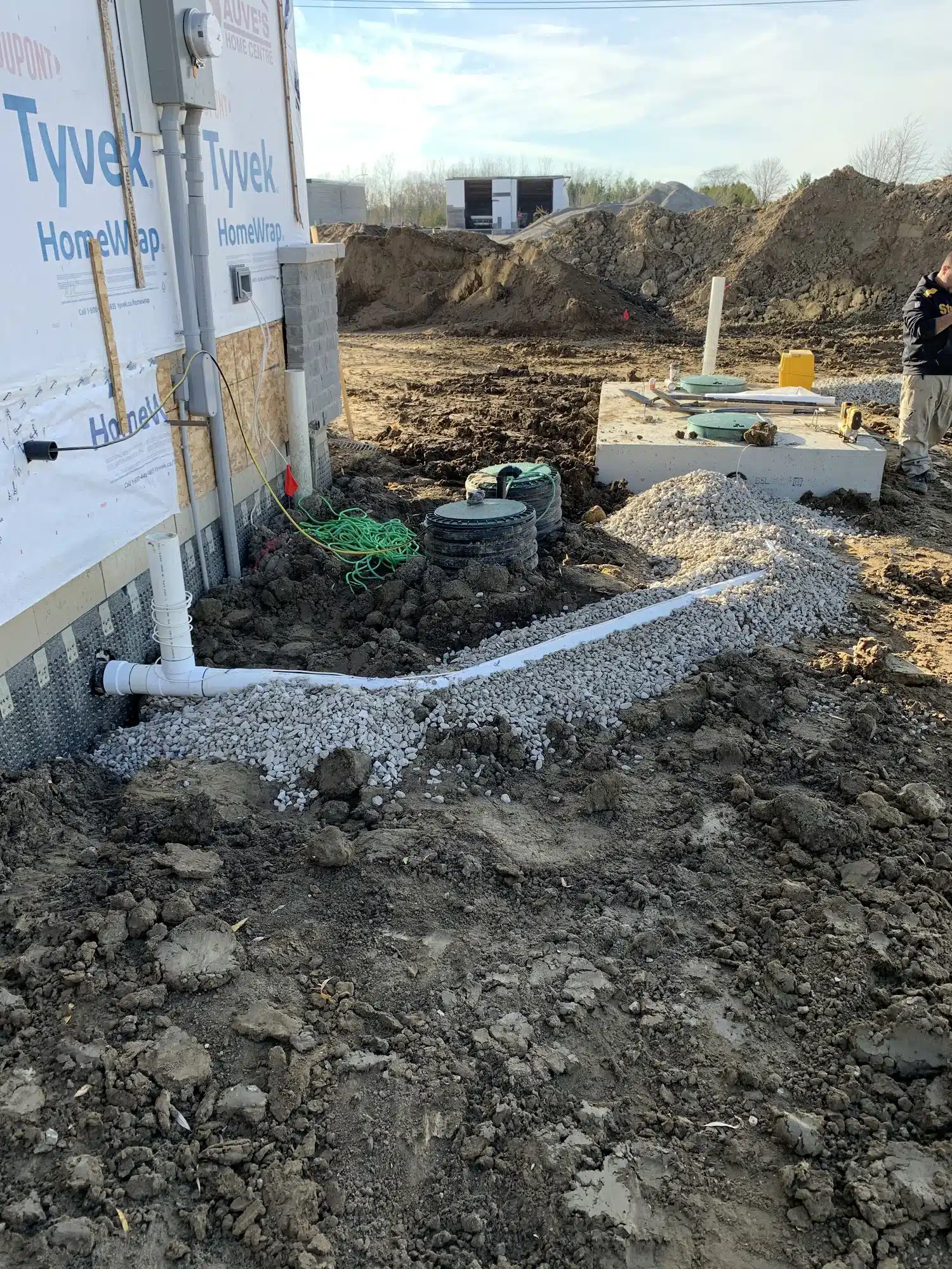 A construction site showing drainage pipes installed beside a building with gravel, Tyvek wrap, electrical boxes, and soil piles in the background.