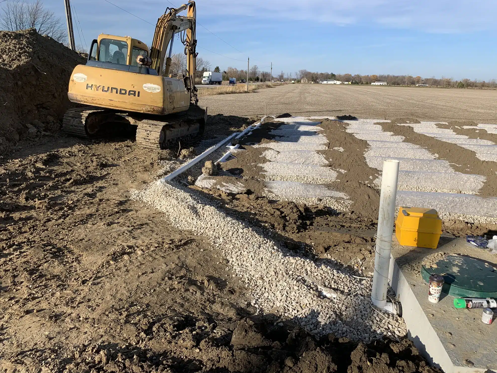 Excavator working near a gravel drainage system installation in a dirt field with construction materials and utility pipes visible.
