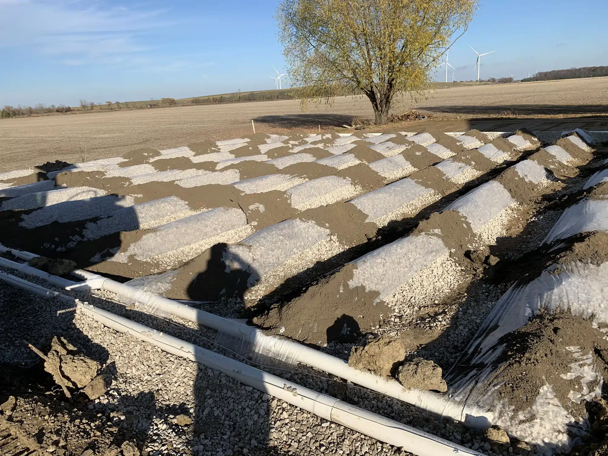 A gravel-covered wastewater leach field with visible white pipes, plastic sheeting, and rows of mounded soil in a rural landscape with a tree and wind turbines in the background.