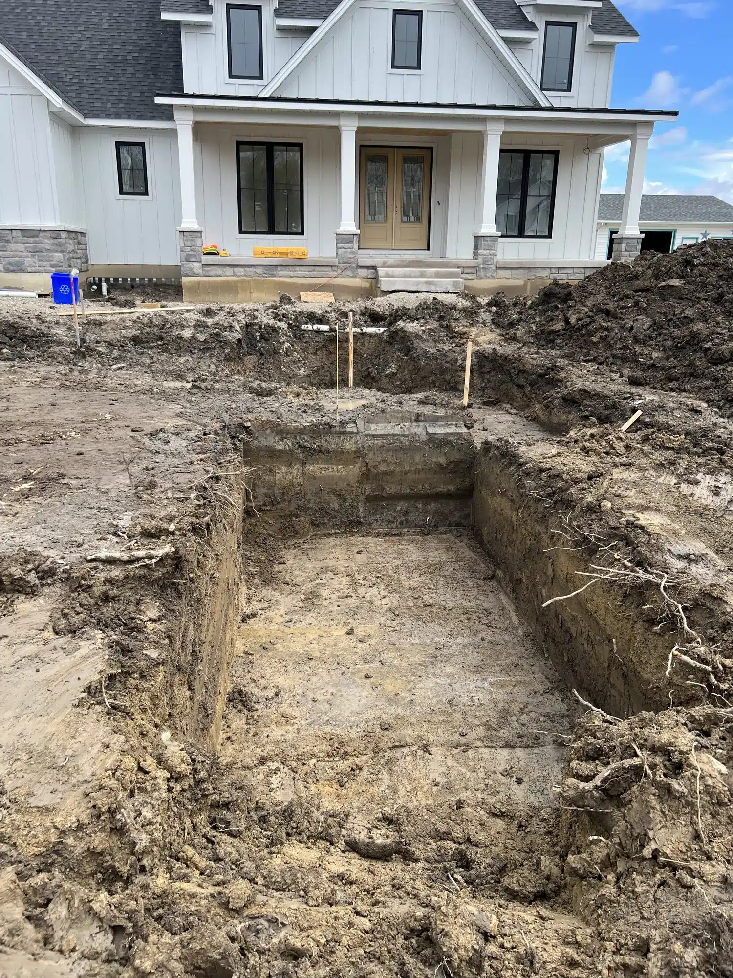 A rectangular hole is dug in front of a house under construction, with dirt piles and building materials visible around the site.