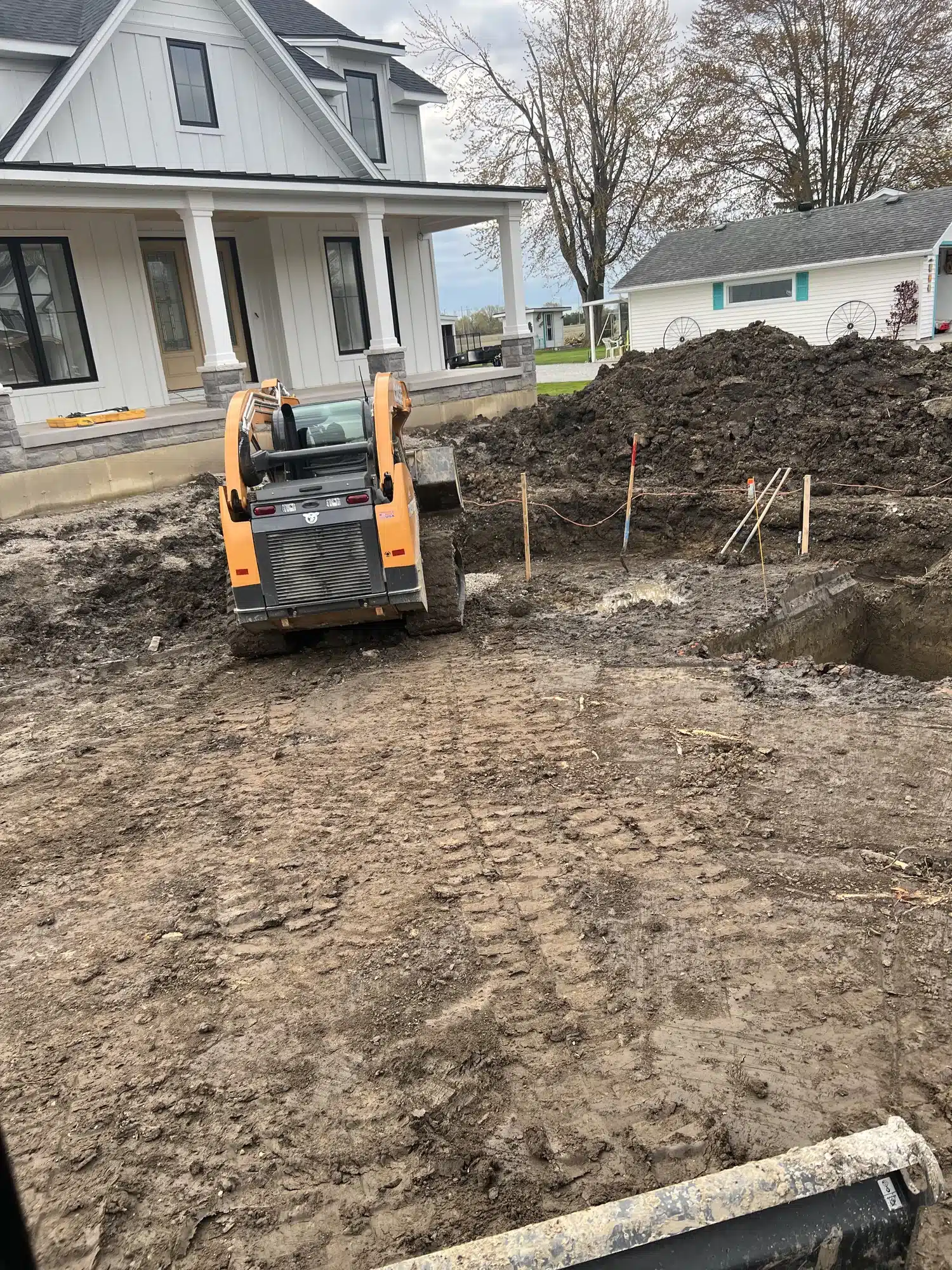 A skid steer loader is parked on dirt near a large hole at a construction site in front of a white house; piles of soil and tools are visible.