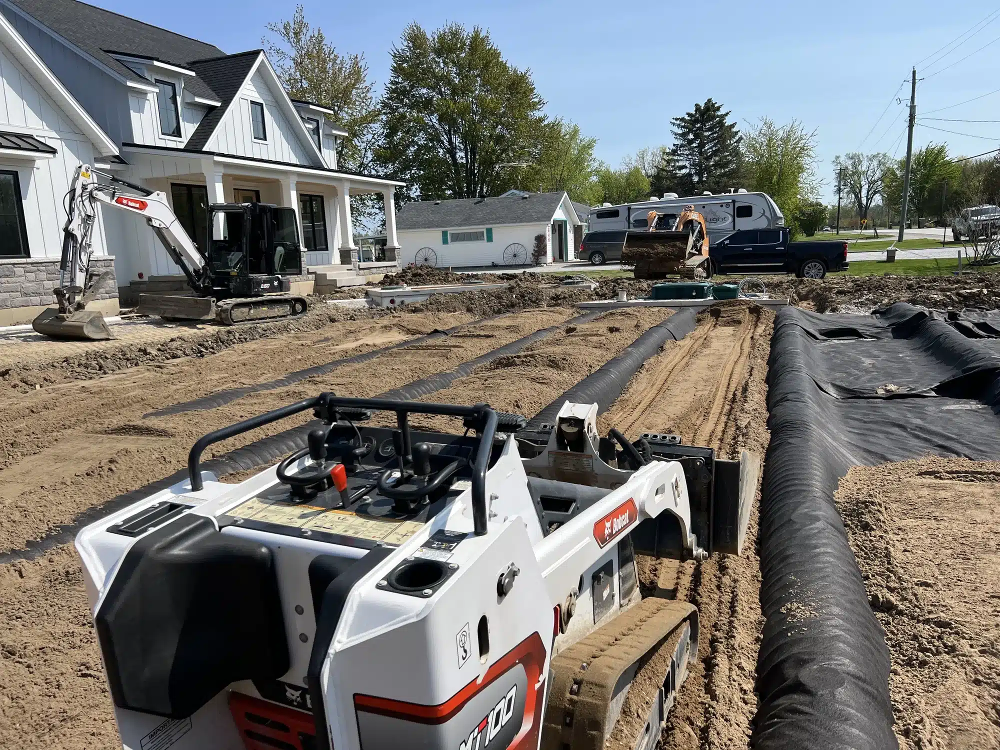 Construction site in front of a house with heavy machinery, including a mini loader and excavator, preparing the ground and laying black liner material.