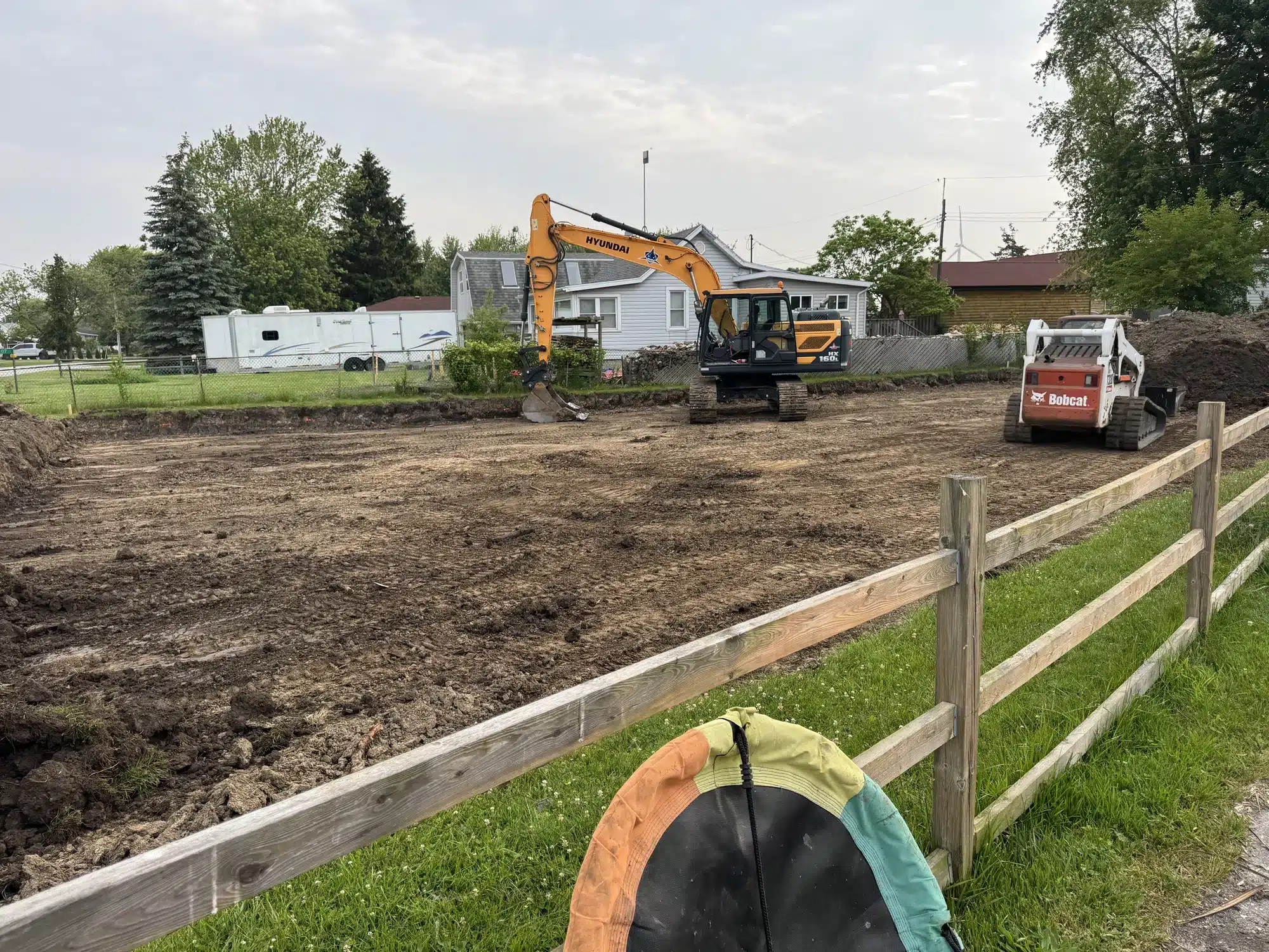 Two excavators, one large and one compact, are clearing and leveling dirt on a fenced lot next to residential houses under a cloudy sky.