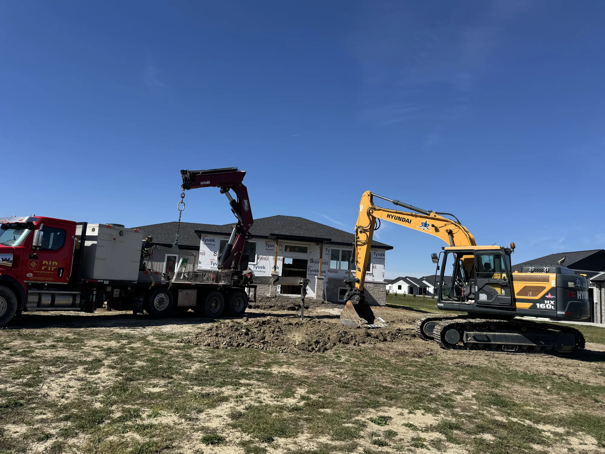 A construction site with a red truck and a yellow excavator working in front of a partially built house under a clear blue sky.