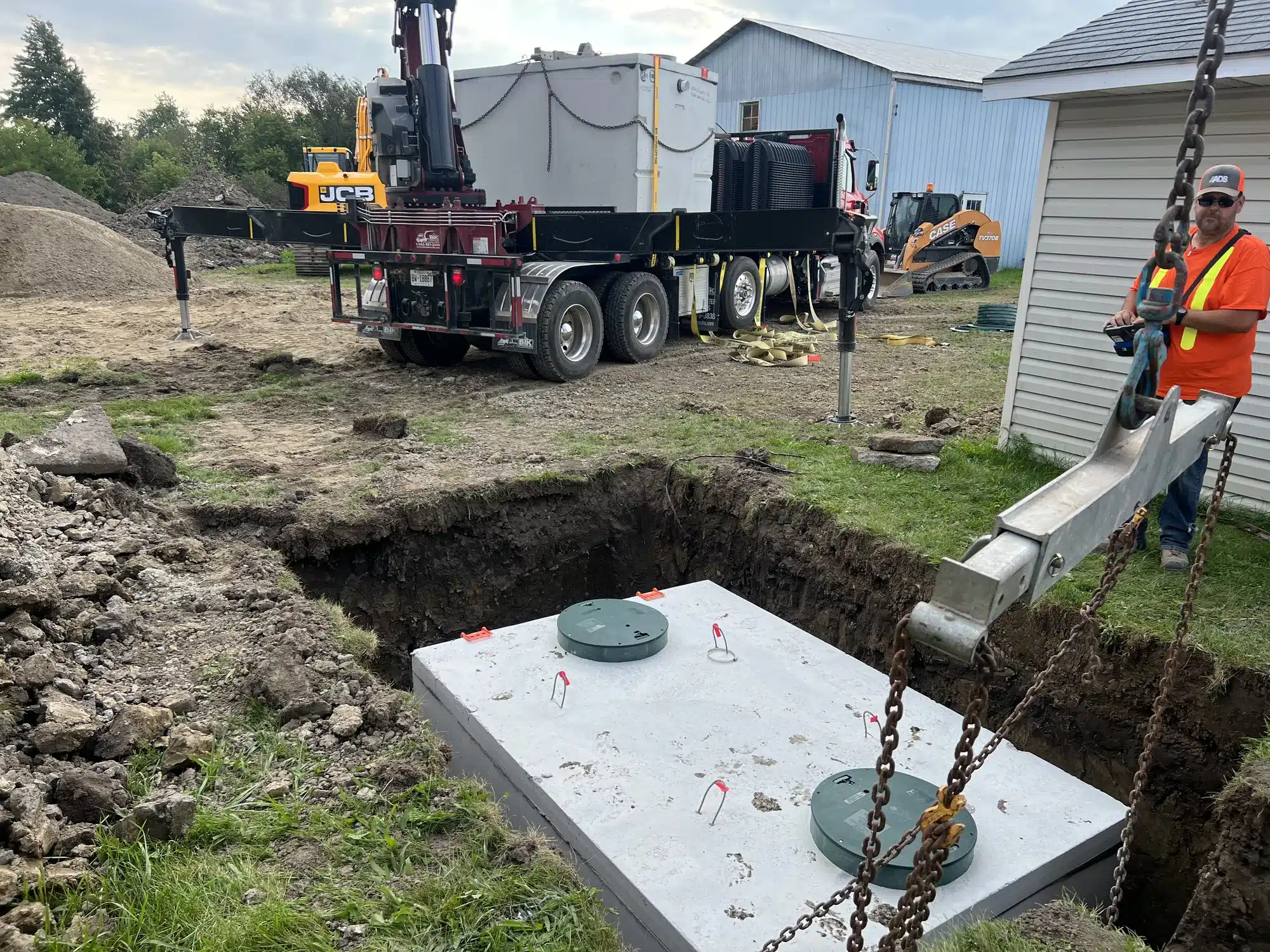 A large concrete septic tank is being installed in a dug-out hole at a construction site, with workers, trucks, and equipment visible in the background.