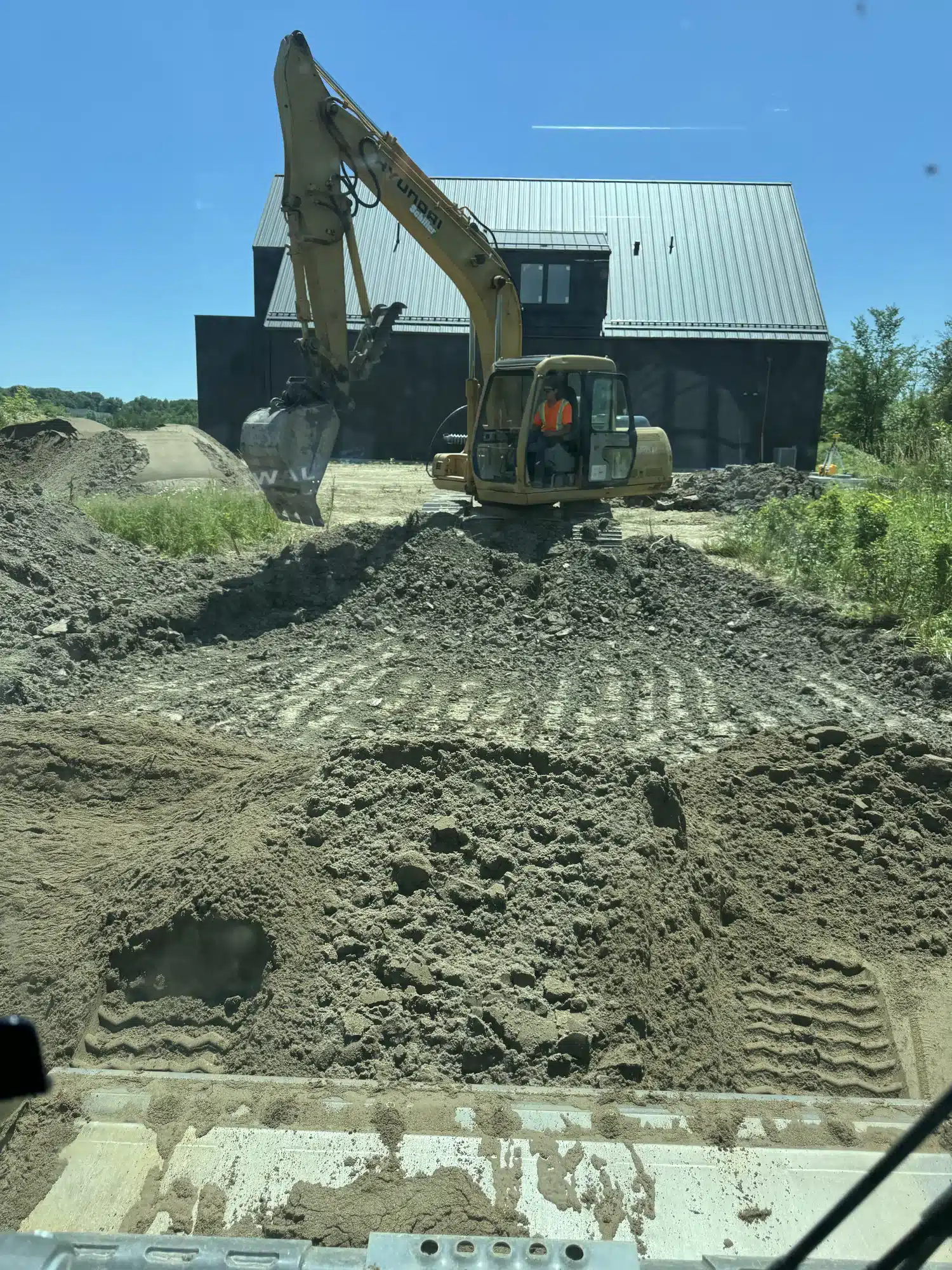 An excavator moves dirt at a construction site in front of a dark building, with a worker visible inside the cab under a clear blue sky.