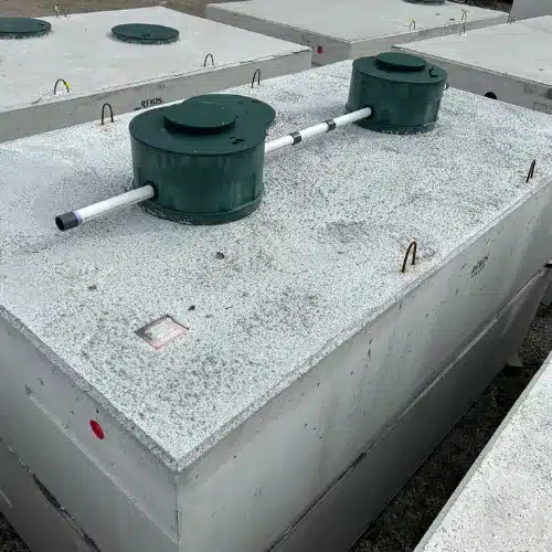 A large rectangular concrete septic tank with two green circular access covers connected by a white pipe, situated outdoors among similar tanks.